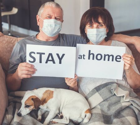 Coronavirus CoVid-19 Couple old aged senior people at home with seasonal winter cold illness disease sit down on the sofa. Elderly couple in medical masks during the pandemicの写真素材
