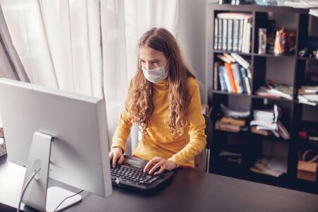 Online distance learning. Teen gaming at home with computer, wearing protective mask. Young girl in quarantine coronavirusの写真素材
