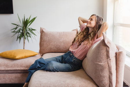 Serene young woman pretty relaxing on couch in living room. Calm happy female freelancer crossing hands behind head, closing eyes and taking a break during remote work day at homeの写真素材