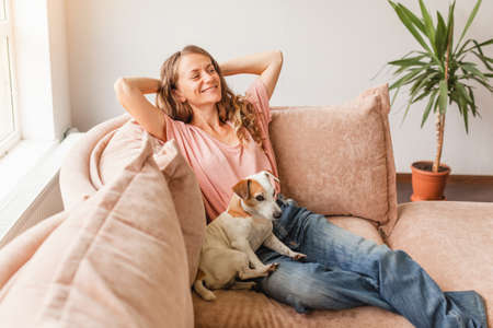 Serene young woman pretty relaxing on couch with her dog in living room. Calm happy female freelancer crossing hands behind head, taking a break during remote work day at homeの写真素材