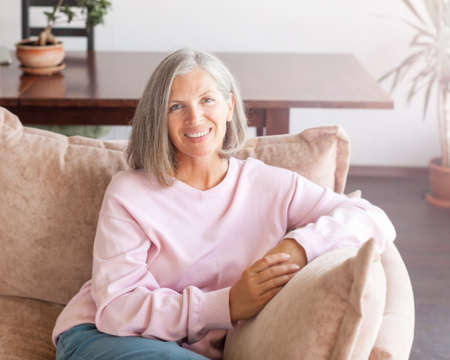 Beautiful middle aged 50 year old female relaxing at home, sitting on comfortable sofa in modern...