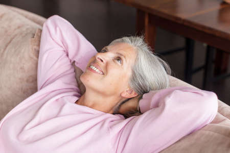In cozy living room happy woman put hands behind head sitting leaned on couch 50s european female enjoy lazy weekend or vacation, housewife relaxing feels satisfied accomplish chores housework conceptの写真素材