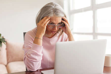Pensive middle-aged woman sit at table in living room using laptop look in distance thinking or pondering, thoughtful senior gray-haired female distracted lost in thoughts feel lonely or sad at homeの写真素材