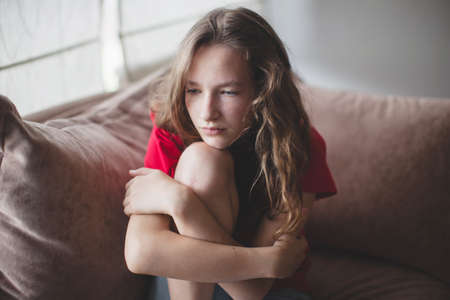 Sad thoughtful teen girl sits on couch feels depressed, offended or lonely, upset young woman suffers from abuse, harassment or heartbreak, grieving lady or violence victim has psychological problemの写真素材