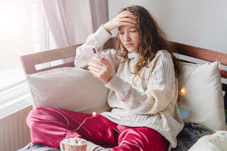Portrait of beautiful young woman with depressed facial expression sitting on gray textile couch holding her phone. Cyber bullying victim concept. Sad female in her roomの写真素材