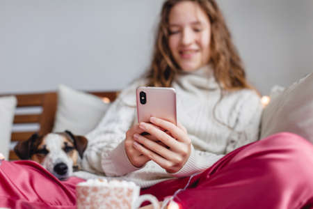 Girl lying on sofa holding phone in her hands in an white sweater on sofa. Teen using cellphone at homeの写真素材