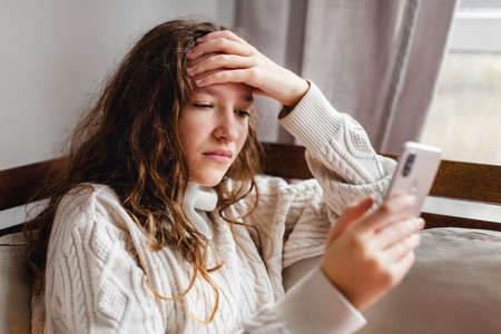 Smartphone Addiction and Depression. Upset teen girl Sitting on couch at home with cellphone. Wooman Looking sadly to device screenの写真素材