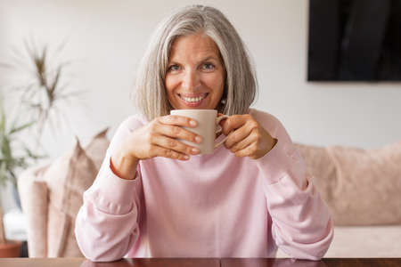 Happy beautiful relaxed mature older adult gray-haired woman drinking coffee relaxing on sofa at home. Smiling stylish middle aged 60s lady enjoying resting sitting on couch in modern living roomの写真素材