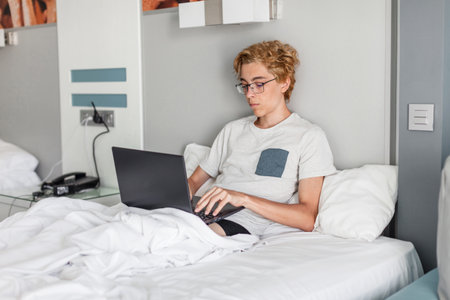 Handsome young man 20s wearing basic gray t-shirt working on laptop pc computer lying in bed with striped sheet pillow blanket resting relaxing spending time in bedroom at home. Teen boy with laptop at campus or hotelの写真素材
