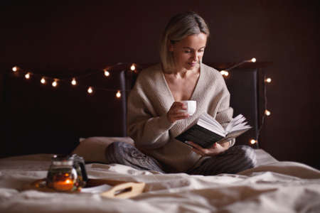 Adult attractive woman is reading book and drinking tea in her bedroom. Middle aged female lies on the bed and holds a book in her handsの写真素材