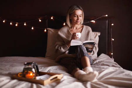 Adult 40s woman is reading a book in bed at cozy home. Middle-aged female is resting in the bedroom on a winter or autumn dayの写真素材
