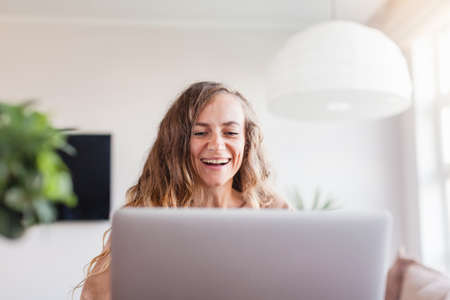Beautiful cheerful woman using silver laptop while sitting on sofa in living room at homeの写真素材