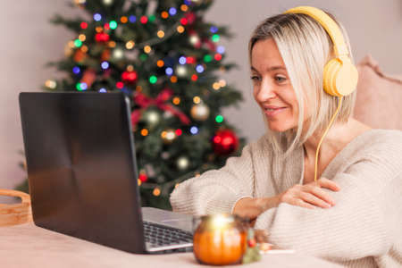 Happy middle-aged woman wearing headphones with a laptop on the background of a Christmas tree. Adult lady communicates online or watch a webinar during the Christmas holidaysの写真素材