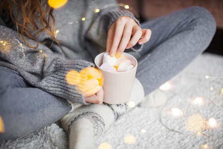 Girl drinks cocoa with marshmallows in a cozy atmosphere. Woman in a warm knitted sweater holds a...