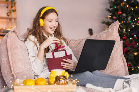 Happy teenage girl is sitting at home on the couch and opening a Christmas gift. Young woman communicates on laptop, make video call and wishes a happy New year and Christmasの写真素材