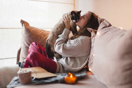 Young beautiful woman kissing cat, sitting at home. Happy girl in sweater in cold weather winter. Female with petの写真素材