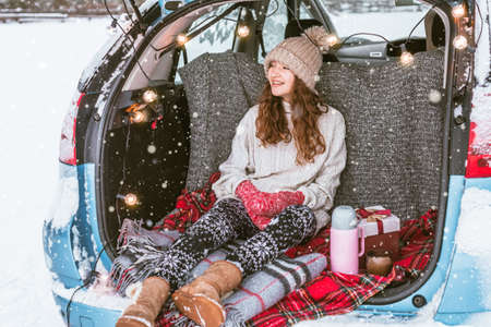 Young woman sitting in car trunk in snowy winter forest. Teen girl relaxing outdoors during road tripの写真素材