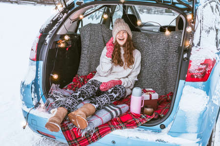 Woman with hot tea sitting in car trunk and looking at the forest, traveling by car during winter holidaysの写真素材