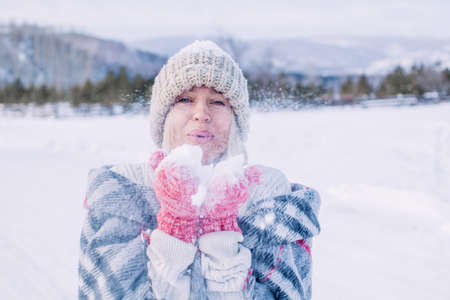 Middle-aged woman walks through the winter forest. Happy female wearing in warm cozy knitted clothes blows on the snow in her palmsの写真素材