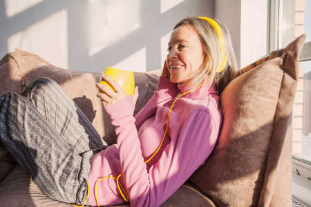 Peaceful middle-aged female in modern headphones sit relax on comfortable couch listening to music,...