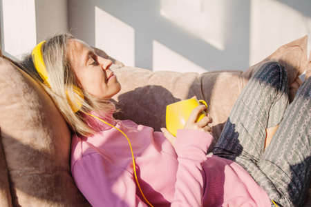 people, technology and leisure concept - happy middle-aged 40s woman sitting on sofa with headphones...