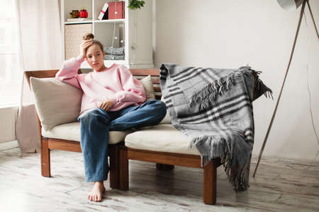 Upset millennial girl sit on couch in living room lost in thoughts thinking pondering of problem solution, depressed unhappy young womanの写真素材