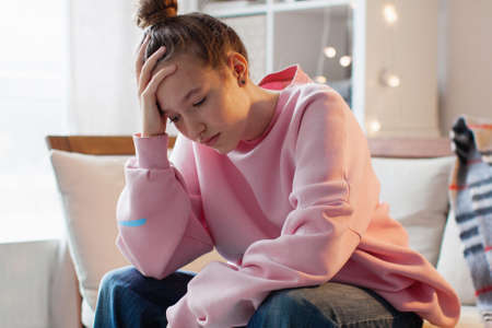 Unhappy depressed teenage girl sit on couch at home suffer from school bullying or discrimination, sad teen outcast feel lonely have difficulties with communication, puberty conceptの写真素材