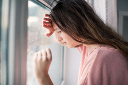 Thoughtful girl looking at window, sad depressed teenager spending time alone at home, young upset pensive woman feeling lonely or frustrated thinking about problemsの写真素材