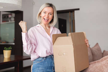 Happy middle aged woman sitting on couch at home opening carton box received parcel package from relatives or shopper make order internet website satisfied client, easy and fast service commerce delivery conceptの写真素材