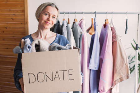 Smiling middle aged woman putting clothing into donation box, sitting on couch at home, happy candid girl packing clothes, Female helping to poor people, charity and aid conceptの写真素材