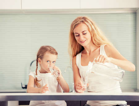 Child with mother drinking water from glass. happy family at home in kitchenの写真素材