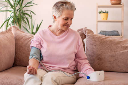 Older elder woman measuring blood pressure, using digital tonometer machine at home. Healthcare, disease treatment concept.の写真素材
