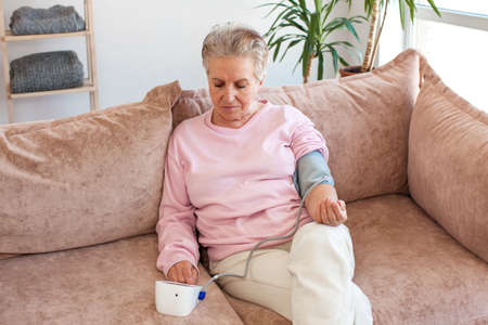 Senior elder woman measure high low blood pressure test holding using medical digital electronic meter sit on sofa at home, heart disease atherosclerosis prevention, hypotension hypertension conceptの写真素材