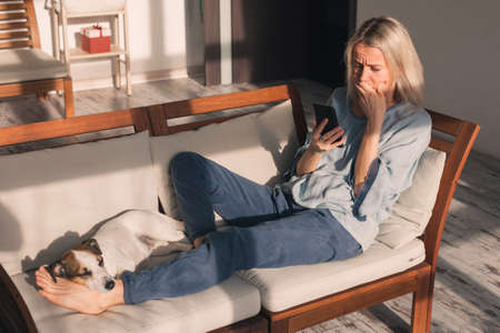 Adult sad woman reads bad news on her phone. Unhappy middle-aged female on couch at homeの写真素材