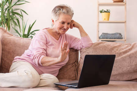 Smiling senior caucasian woman sitting on couch wave talk on video call on laptopの写真素材