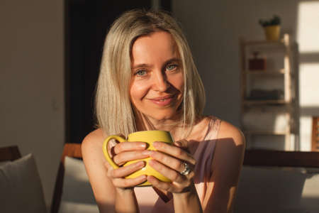 Beautiful middle-aged woman at home sitting on couch in front of window, relaxing in her living room and drinking coffee or teaの写真素材