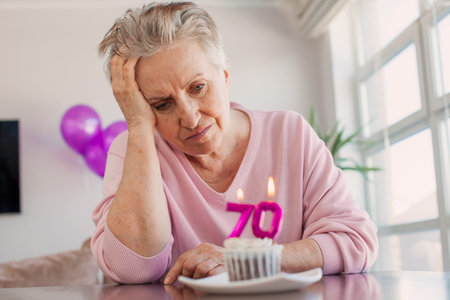 Sad Senior woman holding cake with number 70 and celebrating birthday at homeの写真素材