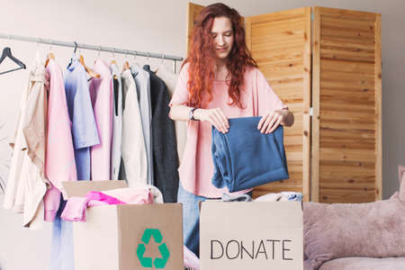 Happy young woman holding cardboard donation box with clothes. Smiling compassionate teen girl preparing staff for charity, reusage recycling concept.の写真素材