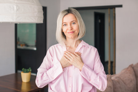 Adult 40s beautiful woman wearing casual shirt standing at home smiling with hands on chest and grateful gesture on face. Health or gratitude conceptの写真素材