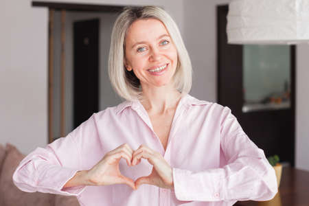 Cute happy middle aged woman making heart shape hand gesture looking at camera, funny smiling adult single woman blogger laughing face showing love sign symbol, dating concept, close up portraitの写真素材