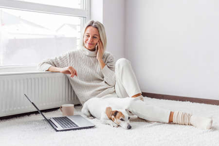 Happy middle aged woman with dog using her laptop and mobile phone at cozy white home. Female freelancer working in living roomの写真素材