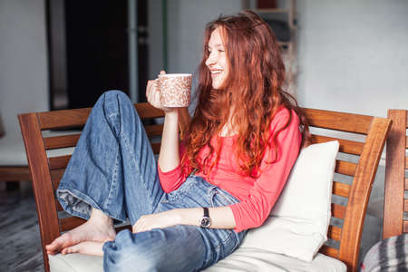 Happy young woman sitting on sofa in cozy clothes with cup of coffee or tea. Smiling teen at cozy home, top viewの写真素材