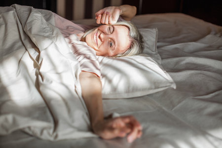 Beautiful middle aged woman waking up in her bed in the bedroom, happy 40s female is stretching and smilingの写真素材