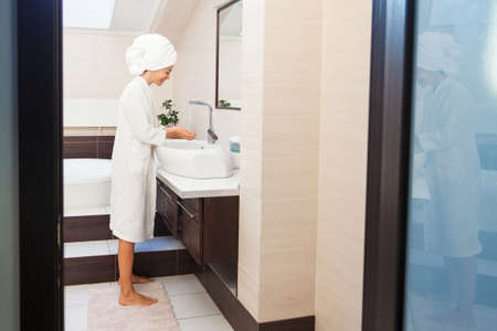 Beautiful middle-aged woman washing face with tap water in bathroom, pretty 40s lady wrap towel on head, skin care treatment conceptの写真素材