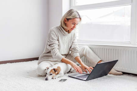 Adult girl is sitting in a bright room and chatting with friends on social networks. Smiling middle-aged woman spends weekend at home with her dogの写真素材