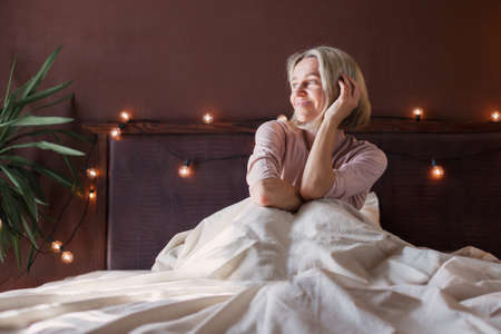 Pretty middle aged woman on bed in modern apartment smiling after wake up. Female in bedroom at homeの写真素材