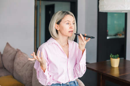 Smiling adult woman relax at cozy home with smartphone, happy middle-aged female sit on couch using cellphone, talking by speakerphone, recording voice message on phoneの写真素材
