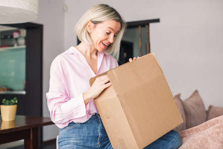 Happy middle aged woman unboxing cardboard box with present inside after buying ordering online via internet a present. Smiling attractive adult female at homeの写真素材