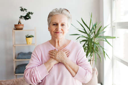 Adult senior 60s beautiful woman wearing casual shirt standing at home smiling with hands on chest...