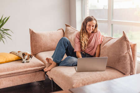 Beautiful cheerful woman using silver laptop while sitting on sofa in living room at homeの写真素材